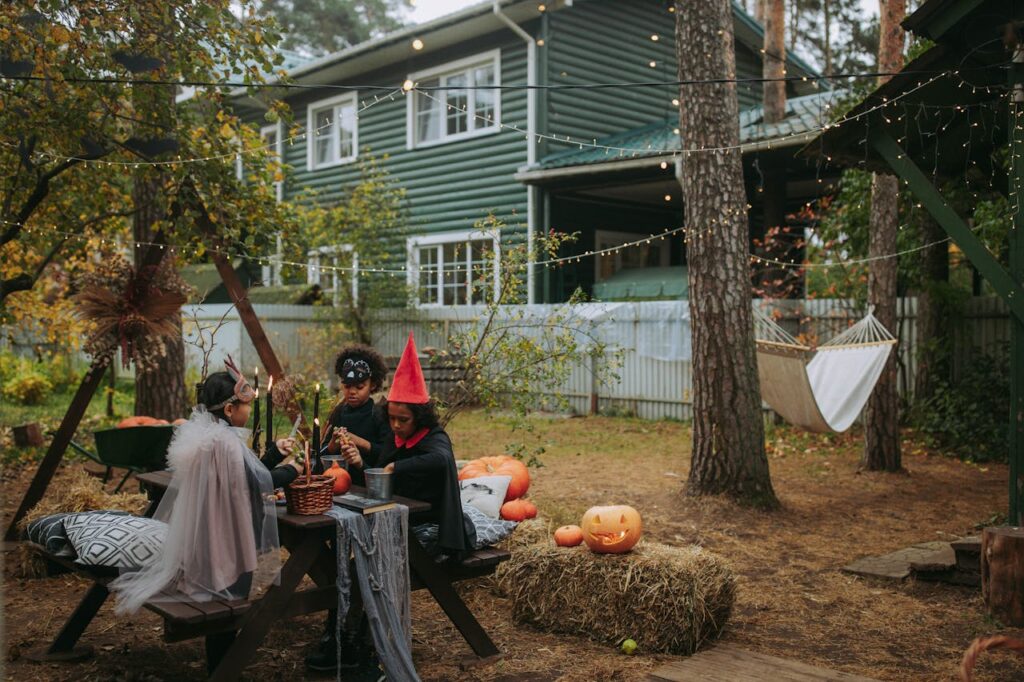 pexels-photo-5607533 Children in Halloween costumes sit outdoors at a picnic table with decorations and string lights.